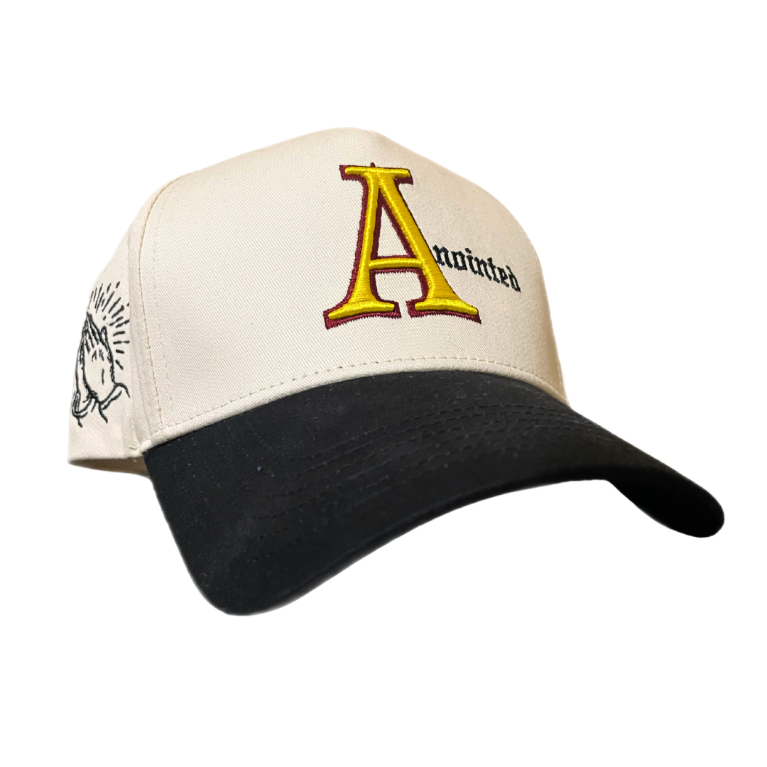 Two-tone baseball cap with a cream crown and black brim. The front of the cap features the word 'Anointed' with a large, stylized red and yellow letter 'A' and black gothic-style text for the remaining letters. The side of the cap displays a small embroidered badge of prayer hands.