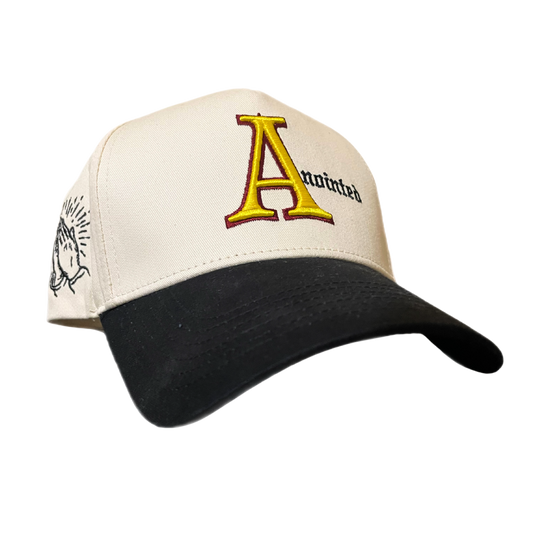 Two-tone baseball cap with a cream crown and black brim. The front of the cap features the word 'Anointed' with a large, stylized red and yellow letter 'A' and black gothic-style text for the remaining letters. The side of the cap displays a small embroidered badge of prayer hands.