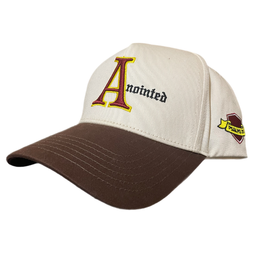 Two-tone baseball cap with a cream crown and brown brim. The front of the cap features the word 'Anointed' with a large, stylized red and yellow letter 'A' and black gothic-style text for the remaining letters. The side of the cap displays a small embroidered badge reading 'PSALMS 23' in yellow text on a maroon and gold ribbon-like background.
