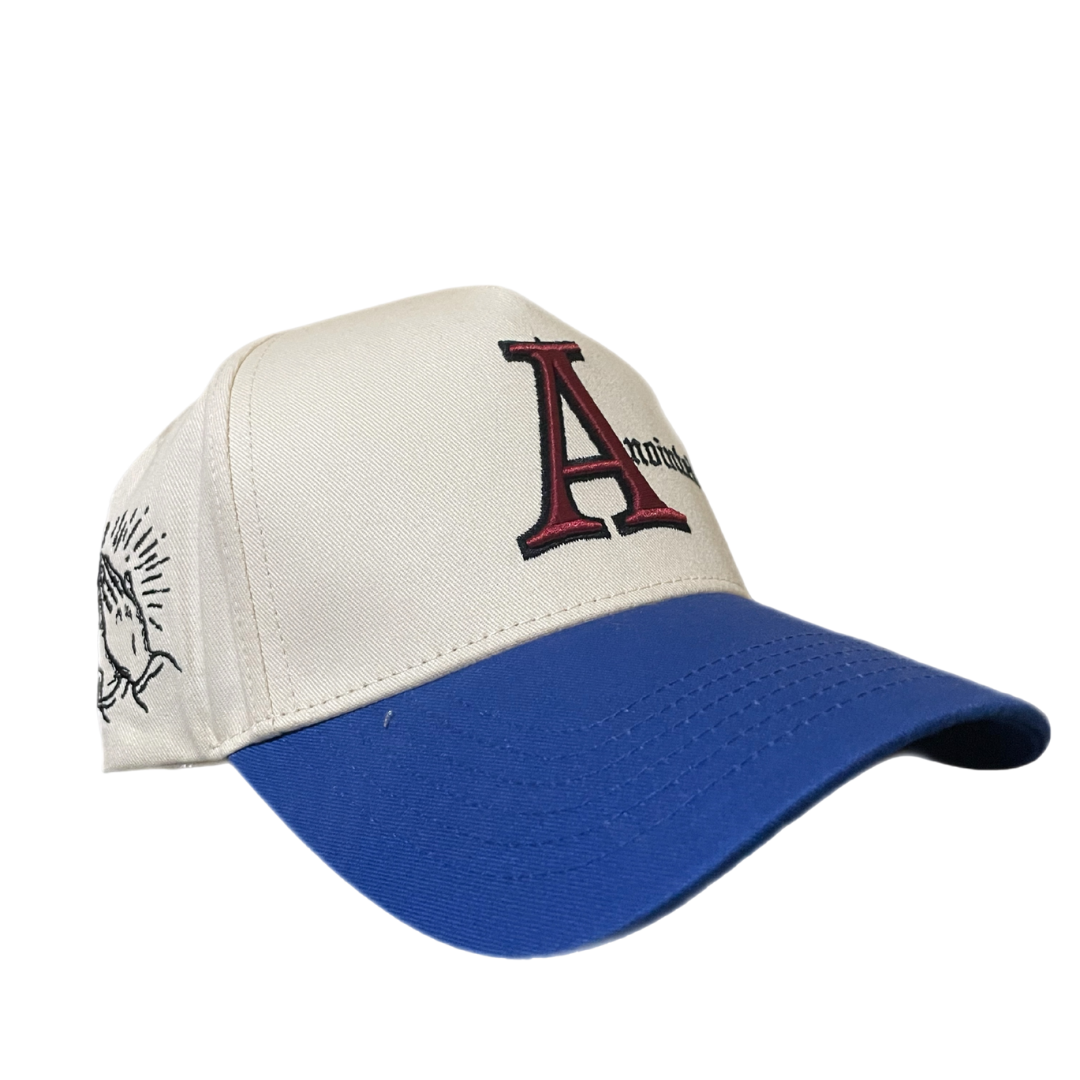 Two-tone baseball cap with a cream crown and blue brim. The front of the cap features the word 'Anointed' with a large, stylized red and black letter 'A' and black gothic-style text for the remaining letters. The side of the cap displays a small embroidered badge of prayer hands.