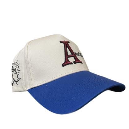 Two-tone baseball cap with a cream crown and blue brim. The front of the cap features the word 'Anointed' with a large, stylized red and black letter 'A' and black gothic-style text for the remaining letters. The side of the cap displays a small embroidered badge of prayer hands.