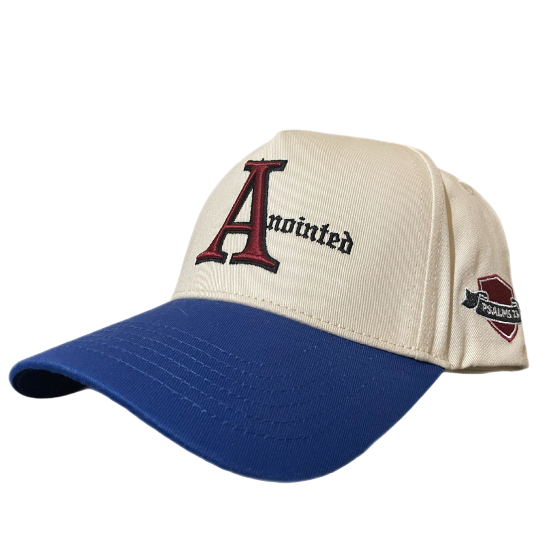 Two-tone baseball cap with a cream crown and blue brim. The front of the cap features the word 'Anointed' with a large, stylized red and black letter 'A' and black gothic-style text for the remaining letters. The side of the cap displays a small embroidered badge reading 'PSALMS 23' in white text on a maroon and black ribbon-like background.