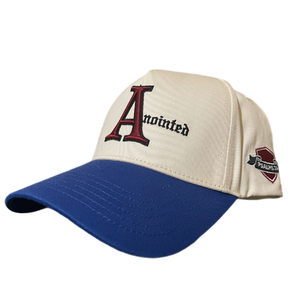 Two-tone baseball cap with a cream crown and blue brim. The front of the cap features the word 'Anointed' with a large, stylized red and black letter 'A' and black gothic-style text for the remaining letters. The side of the cap displays a small embroidered badge reading 'PSALMS 23' in white text on a maroon and black ribbon-like background.