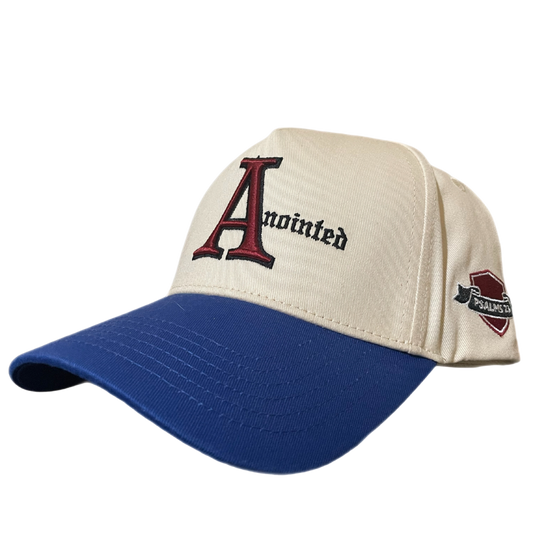 Two-tone baseball cap with a cream crown and blue brim. The front of the cap features the word 'Anointed' with a large, stylized red and black letter 'A' and black gothic-style text for the remaining letters. The side of the cap displays a small embroidered badge reading 'PSALMS 23' in white text on a maroon and black ribbon-like background.
