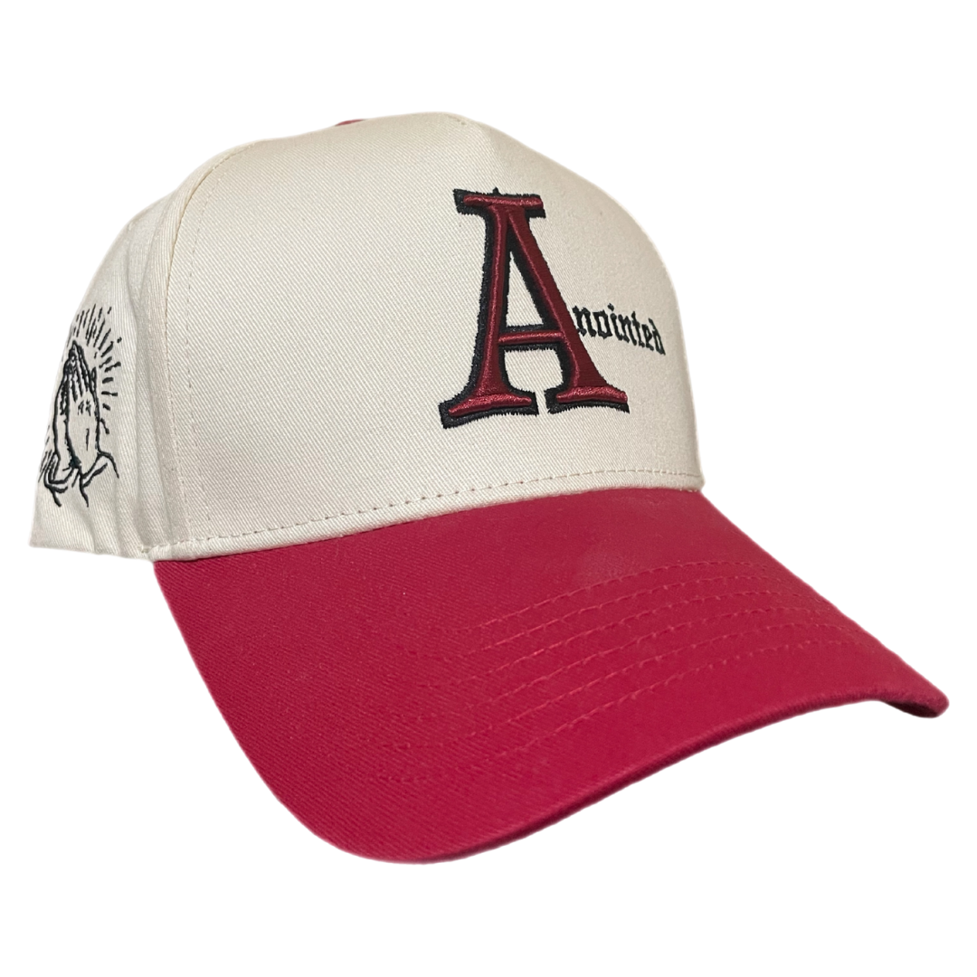 Two-tone baseball cap with a cream crown and red brim. The front of the cap features the word 'Anointed' with a large, stylized red and black letter 'A' and black gothic-style text for the remaining letters. The side of the cap displays a small embroidered badge of prayer hands