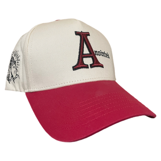 Two-tone baseball cap with a cream crown and red brim. The front of the cap features the word 'Anointed' with a large, stylized red and black letter 'A' and black gothic-style text for the remaining letters. The side of the cap displays a small embroidered badge of prayer hands