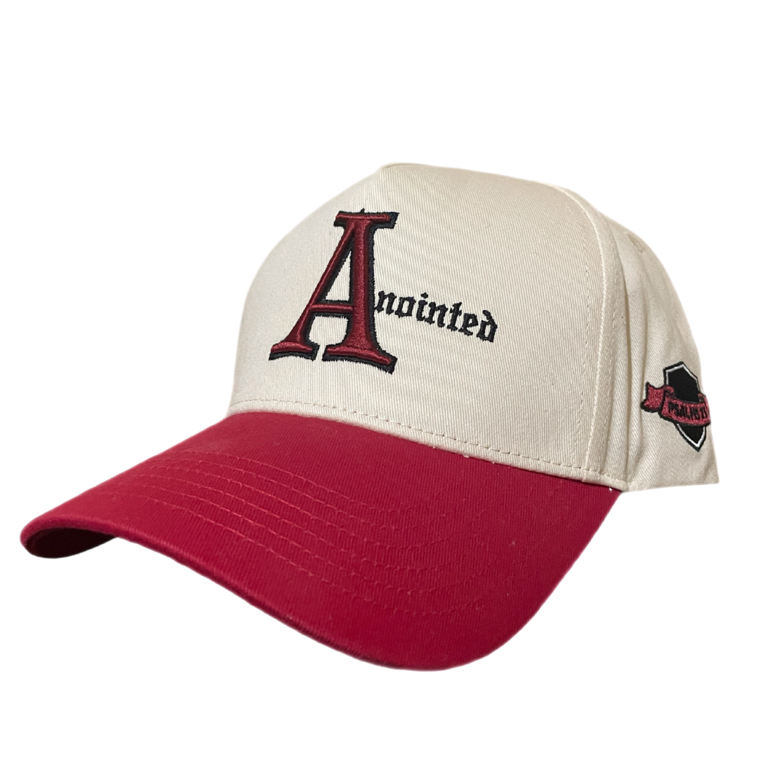 Two-tone baseball cap with a cream crown and red brim. The front of the cap features the word 'Anointed' with a large, stylized red and black letter 'A' and black gothic-style text for the remaining letters. The side of the cap displays a small embroidered badge badge reading 'PSALMS 23' in black text on a maroon ribbon-like background.