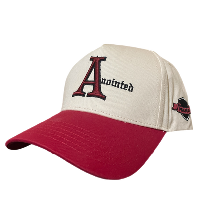 Two-tone baseball cap with a cream crown and red brim. The front of the cap features the word 'Anointed' with a large, stylized red and black letter 'A' and black gothic-style text for the remaining letters. The side of the cap displays a small embroidered badge badge reading 'PSALMS 23' in black text on a maroon ribbon-like background.